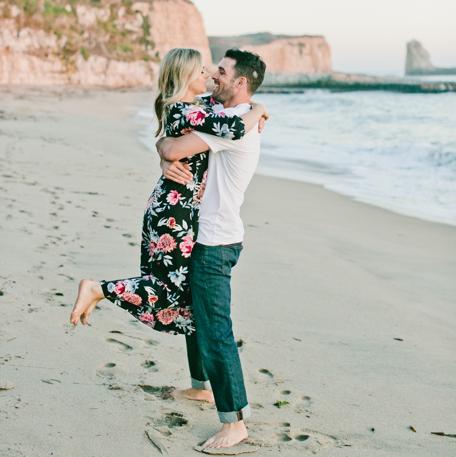 A man lifts up a woman on the beach at sunset.