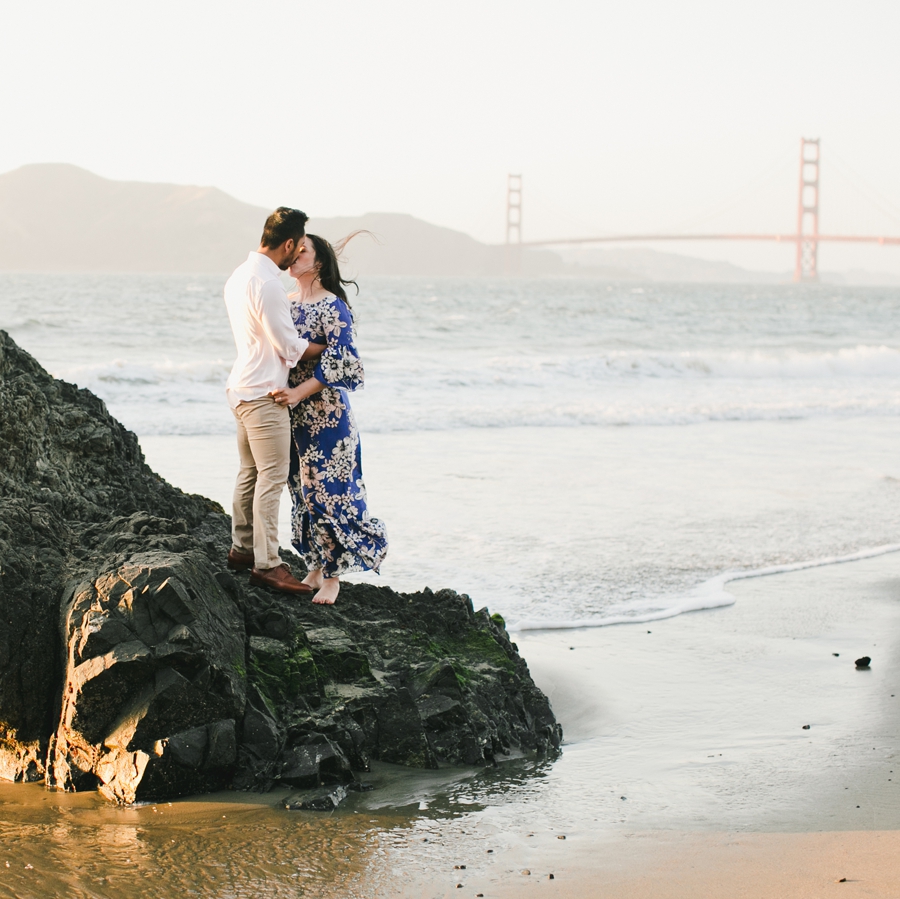 An engagement photo of a couple on a rock at sunset with golden gate bridge in the view.