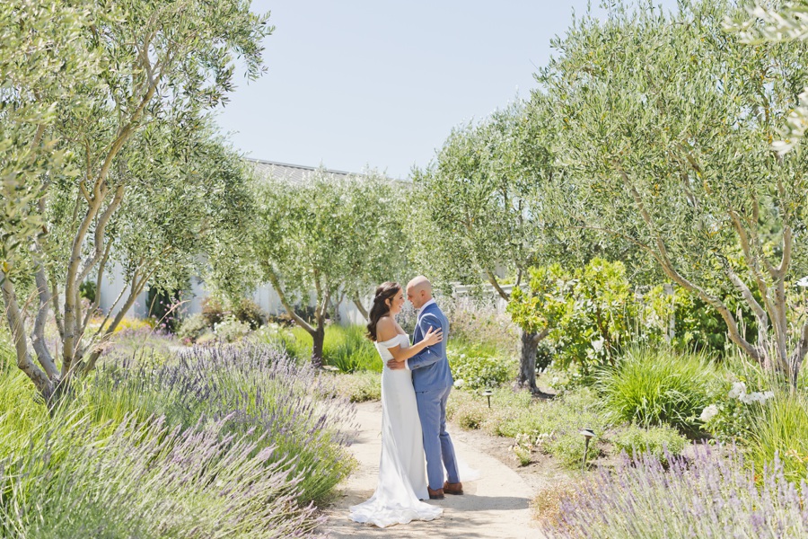A couple in wedding attire embraces on a sunlit garden path surrounded by trees and lavender plants at The Barn at Harrow Cellars.