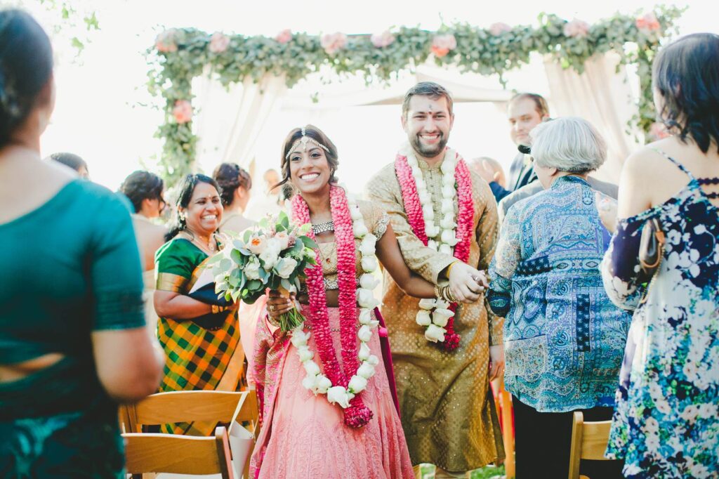 A smiling bride and groom in traditional Indian attire and flower garlands walk down the aisle, surrounded by guests—a joyful moment beautifully captured, reflecting the artistry and value of Bay Area wedding photographer cost.