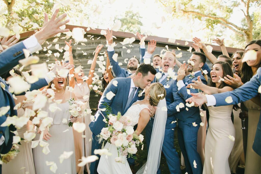 A bride and groom kiss while surrounded by their wedding party, who are tossing flower petals in celebration outdoors—captured perfectly to show the value of Bay Area wedding photographer cost.