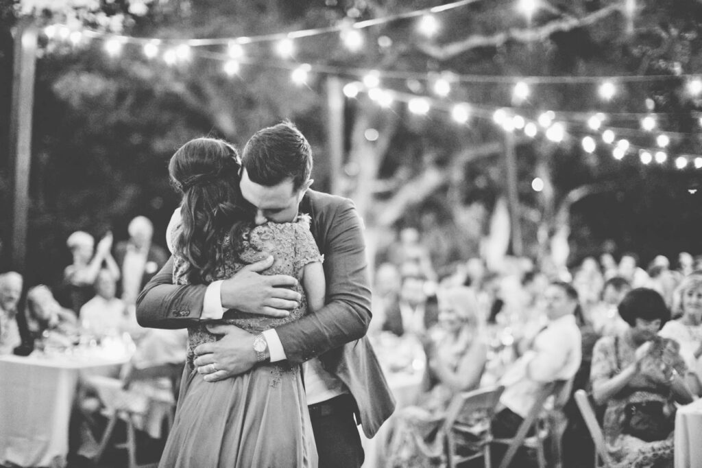 A man and woman embrace tightly at an outdoor event with string lights overhead and seated guests in the background, capturing a moment often highlighted by Bay Area wedding photographer cost discussions.