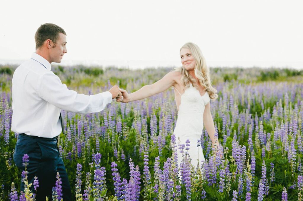 A man and a woman in formal attire hold hands in a field of purple flowers, with the woman in a white dress gazing at the man—capturing moments like this, discover Bay Area wedding photographer cost for your special day.