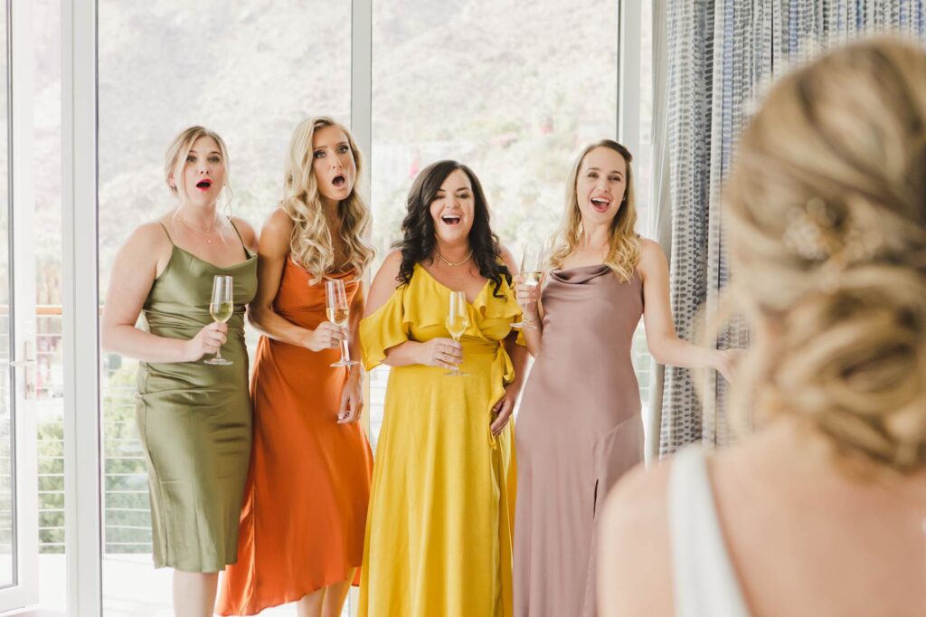 Four women in colorful dresses hold champagne glasses, reacting with surprise and excitement toward a woman in a white dress—capturing the joy that makes discovering Bay Area wedding photographer cost worthwhile.
