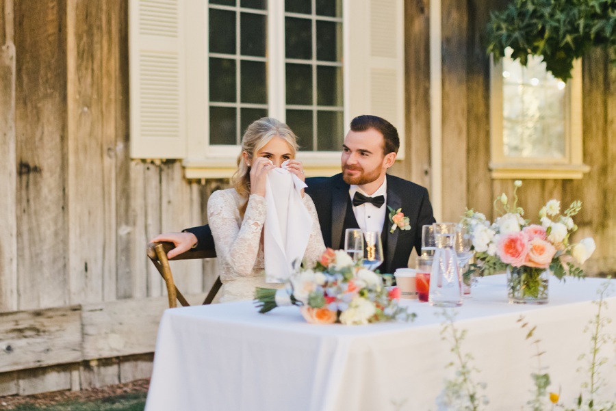 A bride wipes her face with a napkin while sitting beside a groom at a decorated table outdoors, in front of a wooden building with windows.