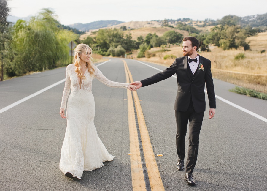 A bride in a white dress and a groom in a black suit hold hands while walking on a rural road with hills and trees in the background.