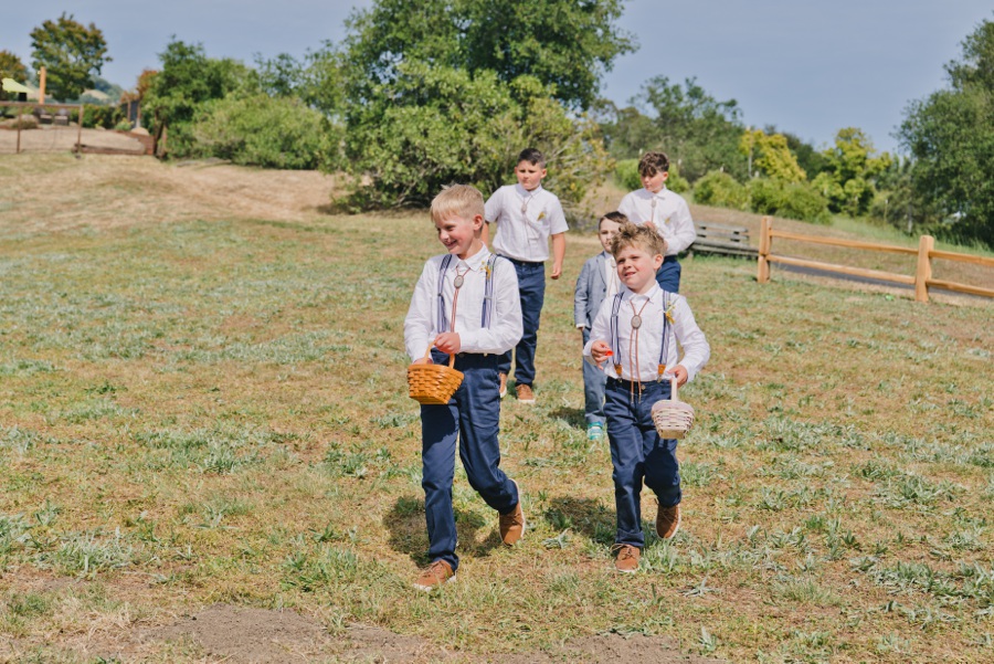 Five young flower boys wearing suspenders and bowties walk across a grassy field, each carrying a small basket—captured in perfect detail by a film wedding photographer from the Bay Area. Trees and a wooden fence are visible in the background.