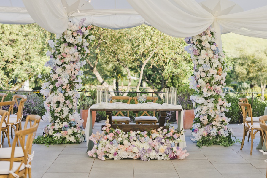 A decorated outdoor wedding table with wooden chairs, surrounded by floral arrangements and draped white fabric under a canopy, with greenery in the background.