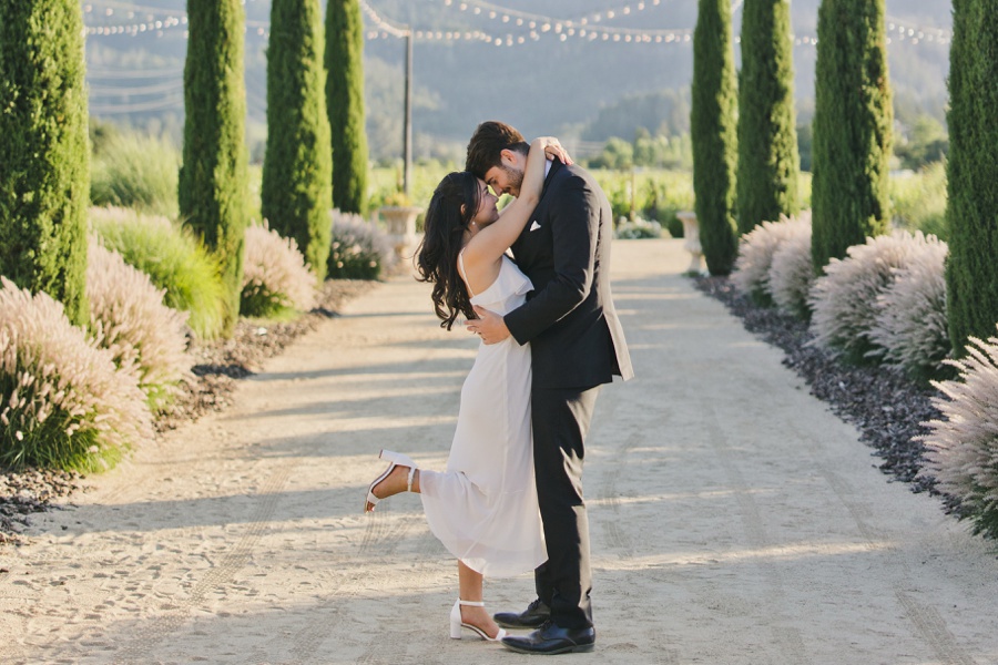 A couple dressed in wedding attire shares an embrace and kiss on a tree-lined, sunlit path with mountains in the background.