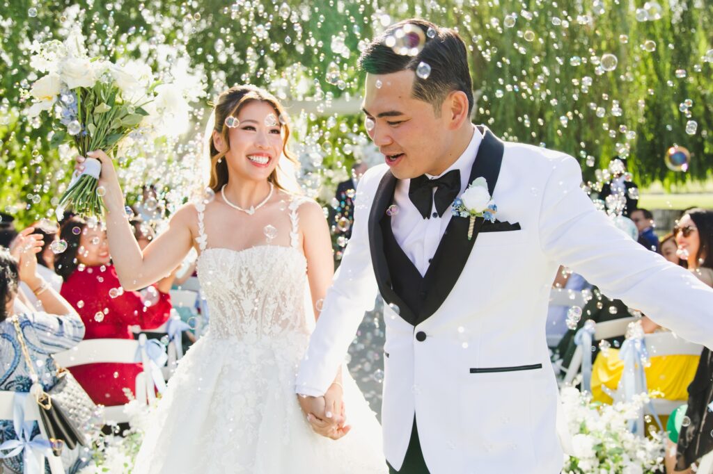 A bride and groom, holding hands and smiling, walk outside among guests as bubbles float in the air—capturing a magical moment for their wedding album.