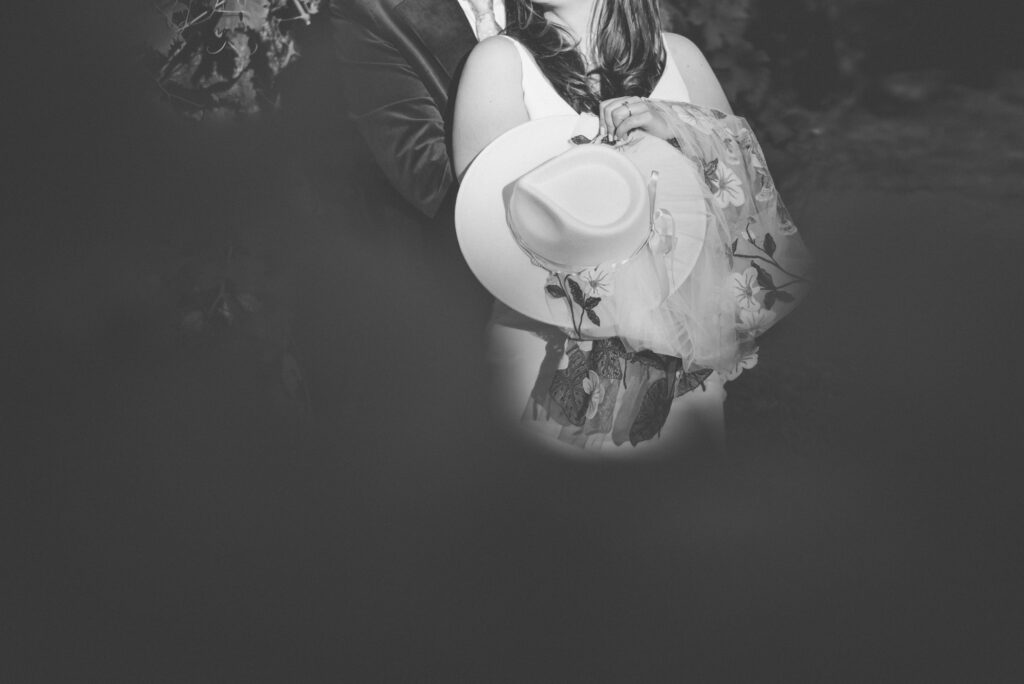 A black and white photo shows a person holding a wide-brimmed hat with a floral scarf, partially blocking their face, while another person stands close behind them.
