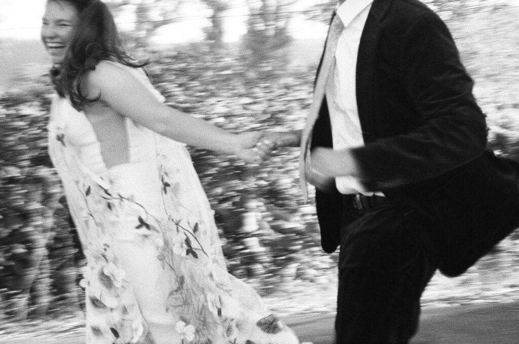 A woman in a floral dress and a man in a suit hold hands and run outdoors, both appearing joyful captured in black and white film. The background is blurred, suggesting motion.