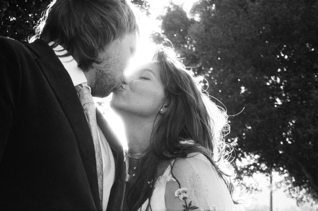 A man and woman share a kiss outdoors in sunlight, both dressed in formal attire, with trees visible in the background captured in black and white film.