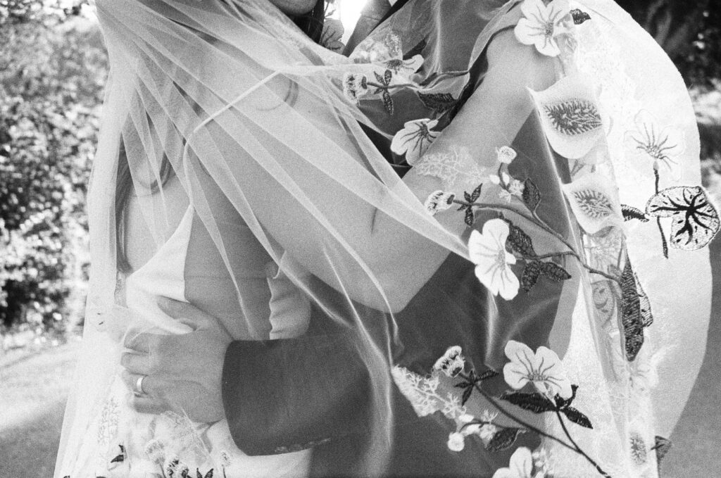 A couple embraces closely, their faces partially obscured by a floral-patterned wedding veil in an outdoor setting captured in black and white film.