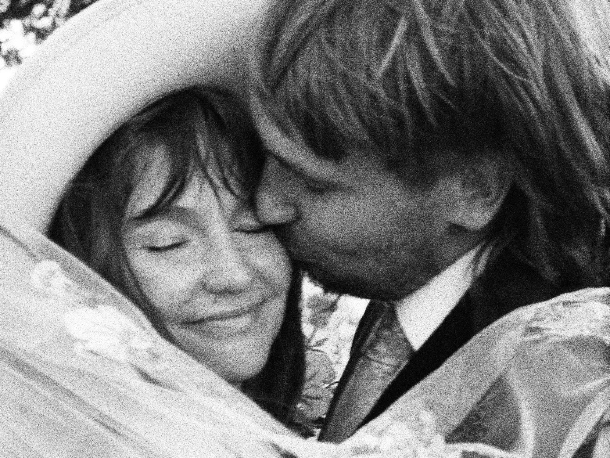 A person with long hair and a suit kisses another smiling person on the cheek; both appear happy and close together, with a veil and hat visible.