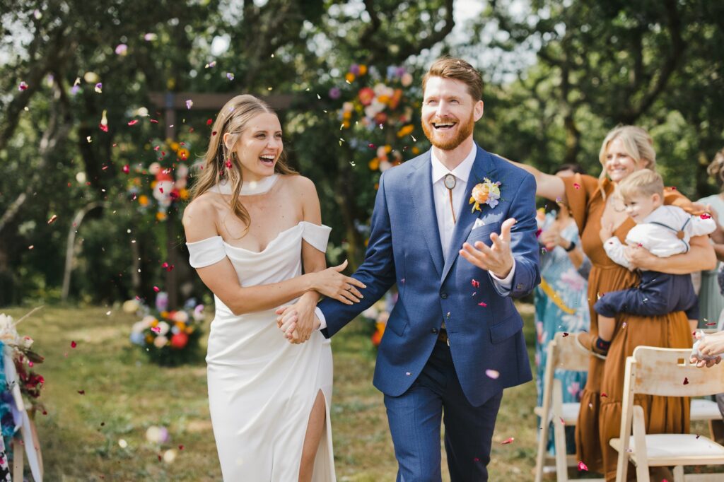 A newlywed couple walks outdoors, smiling and holding hands, as guests throw flower petals—the perfect moment for a wedding album. The bride wears a white dress and the groom wears a blue suit.
