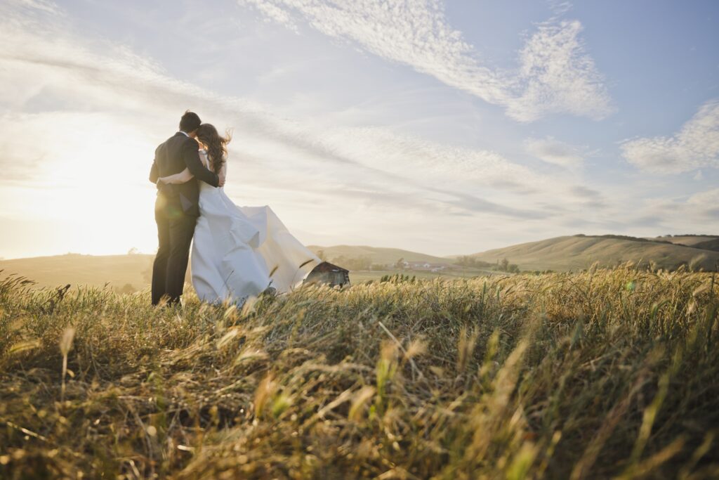 A bride and groom stand together in a field at sunset, embracing as they look toward rolling hills under scattered clouds—a perfect moment for their wedding album.