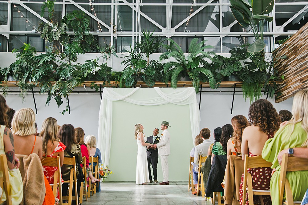 A couple stands with an officiant under a white arbor, exchanging vows in front of seated guests at an indoor wedding with abundant greenery.