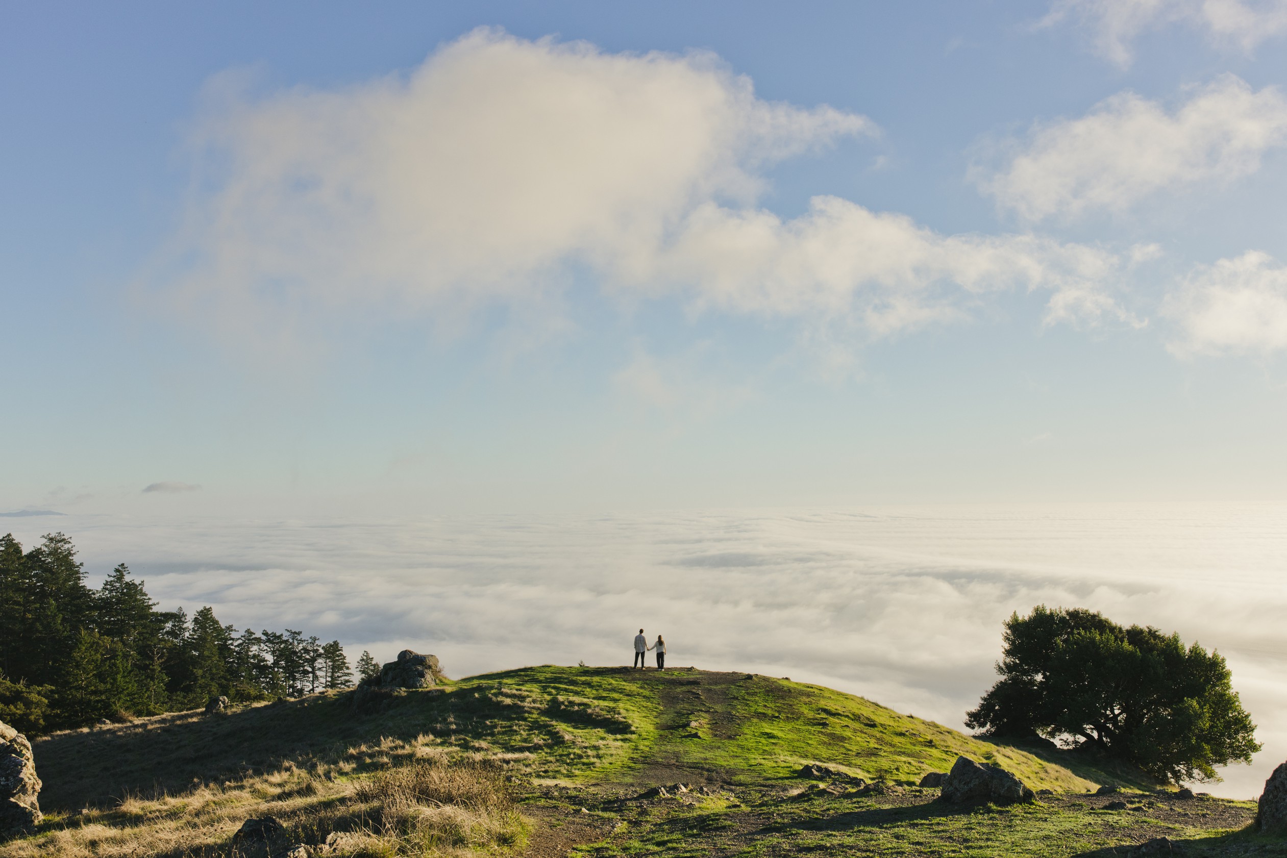 Two people stand on a grassy hilltop overlooking a sea of clouds under a partly cloudy sky, with trees and rocky terrain nearby.