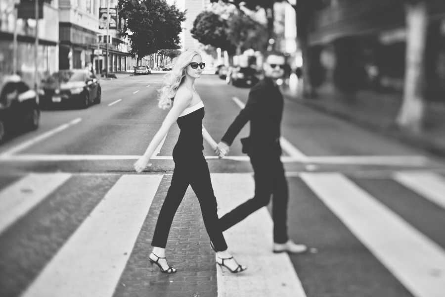 A woman and a man, both in dark clothing, walk hand in hand across a city crosswalk. The image is in black and white and slightly out of focus in the background.