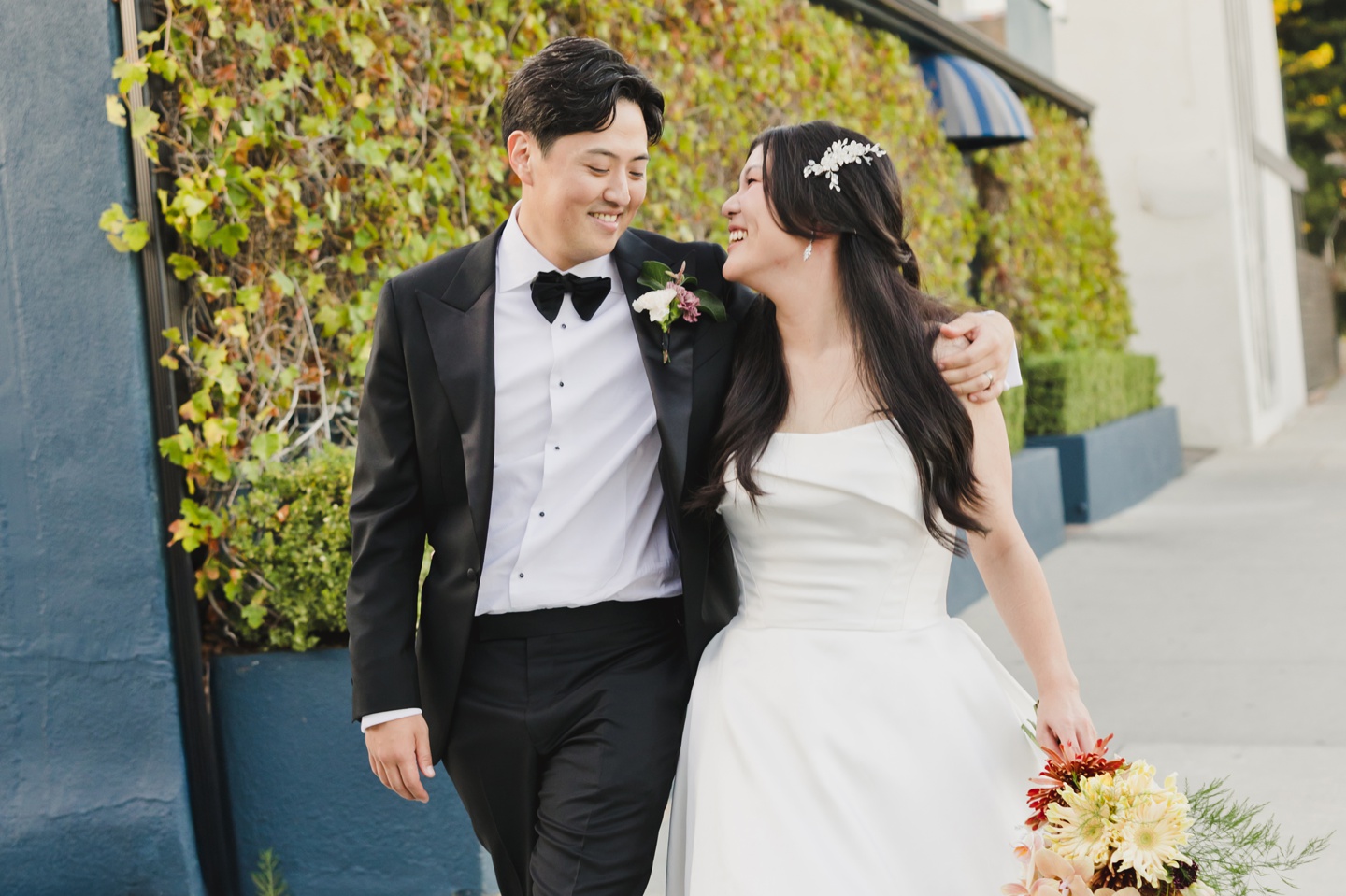 A couple dressed in wedding attire walks outdoors at their Fig House Wedding, smiling at each other. The groom wears a black suit and bow tie; the bride holds a bouquet and wears a white dress with a hair accessory.
