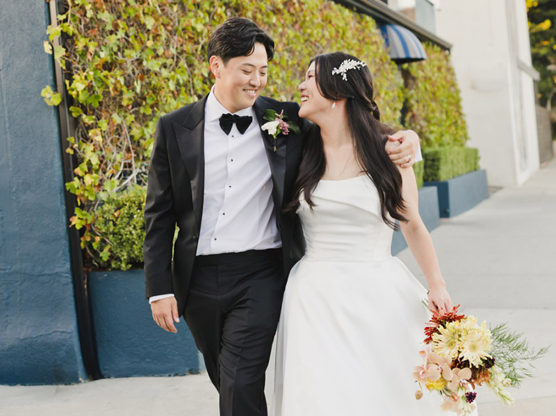A bride in a white dress and a groom in a black tuxedo walk arm in arm outside the Fig House Wedding venue, smiling at each other. The bride holds a bouquet of flowers.