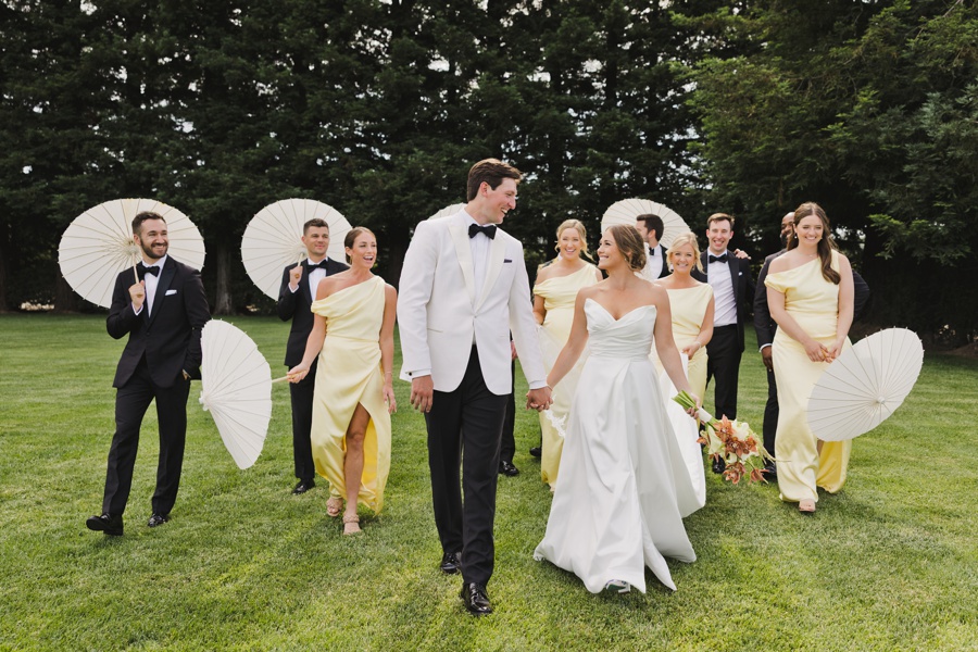 A bride and groom walk on grass, holding hands, surrounded by their wedding party in formal attire, with some holding white parasols. Trees are in the background.