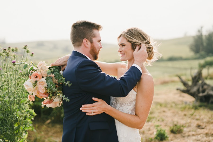 A bride and groom stand outdoors embracing at their Olympia's Valley Estate Wedding, with the groom touching the bride's hair. She holds a bouquet of flowers, set against a backdrop of grassy fields and trees.