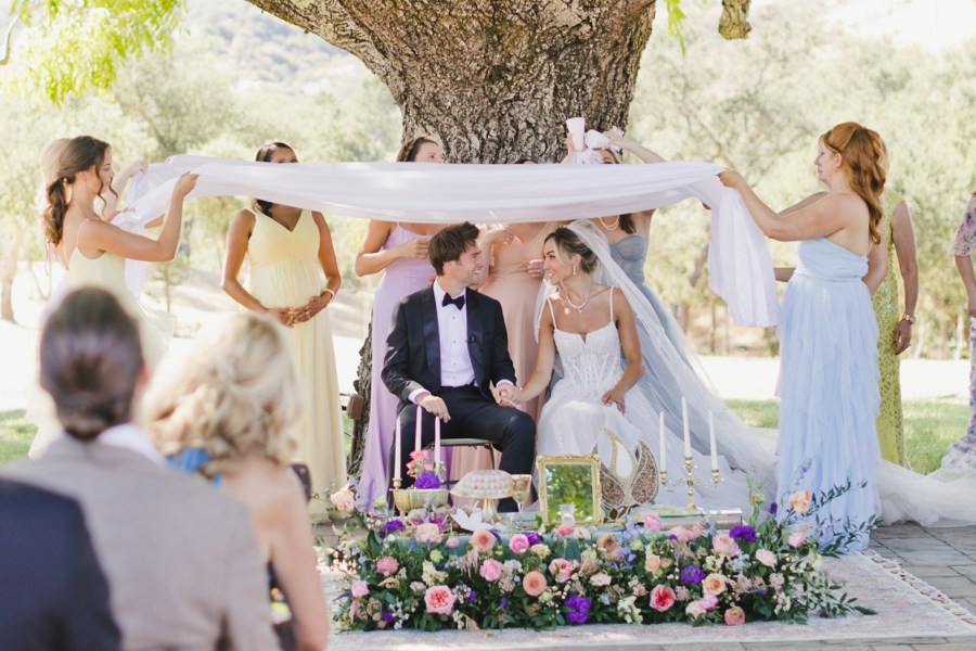 A bride and groom sit under a tree at their SSS Ranch Wedding, surrounded by women holding a white cloth above them, with flowers and candles beautifully arranged in front of them in a stunning outdoor setting.