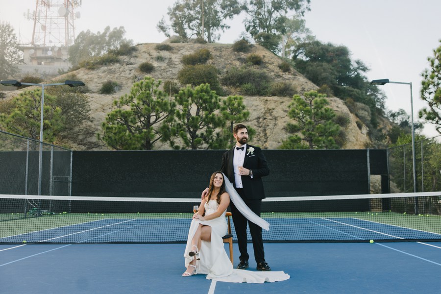 A bride sits on a chair and a groom stands beside her on a tennis court, with hills and trees in the background.