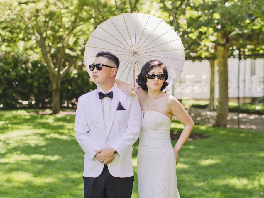 A couple dressed in formal wedding attire and sunglasses stands on grass, with the woman holding a white parasol. Trees and sunlight are visible in the background.