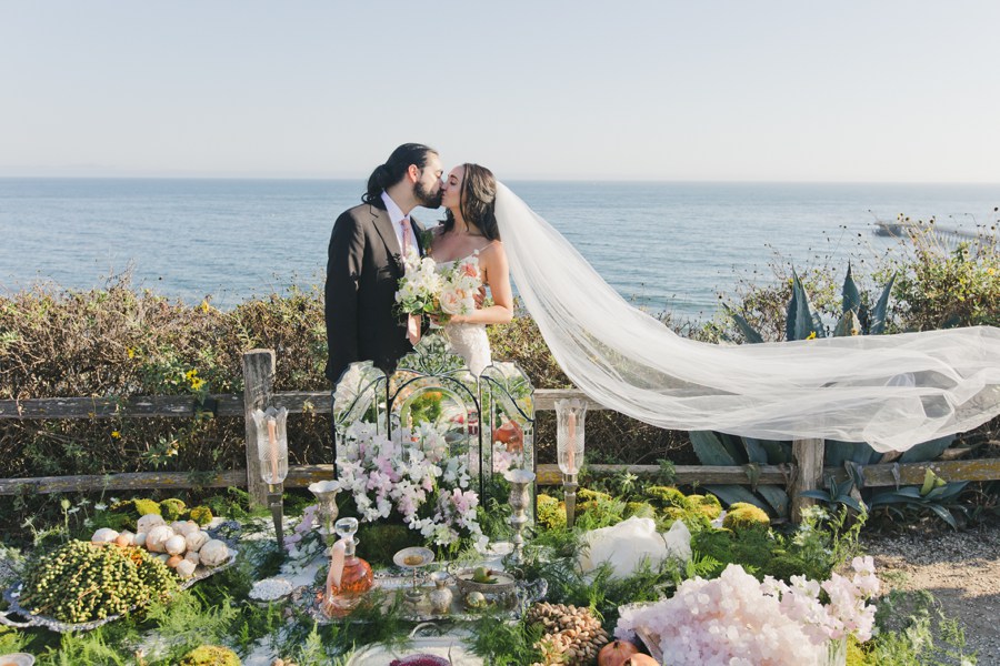 A bride and groom kiss by the ocean, standing behind a decorated Sofreh Aghd at the Bacara Santa Barbara. The bride’s long veil flows in the breeze.