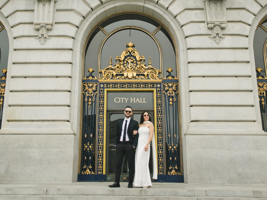 A man in a suit and a woman in a white dress stand arm-in-arm before ornate doors labeled City Hall, capturing the elegance of their San Francisco City Hall engagement session.