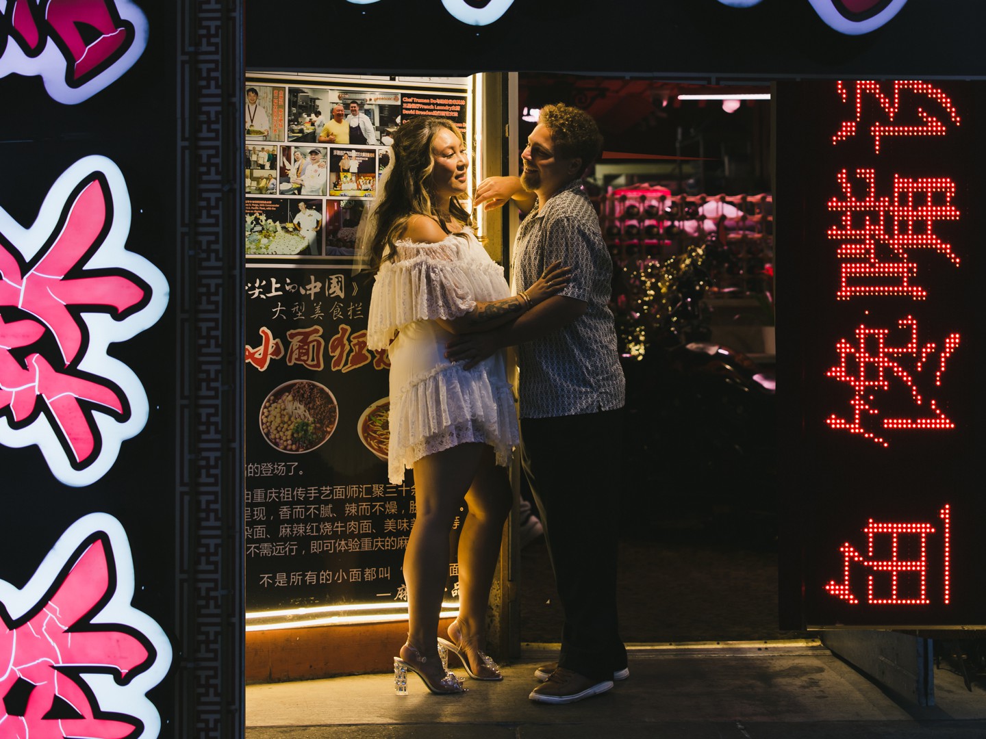 A woman in a white dress and a man stand closely together, smiling, in the entrance of a restaurant in Chinatown with neon signs and food photos on display—a charming moment captured among San Francisco Chinatown engagement photos.