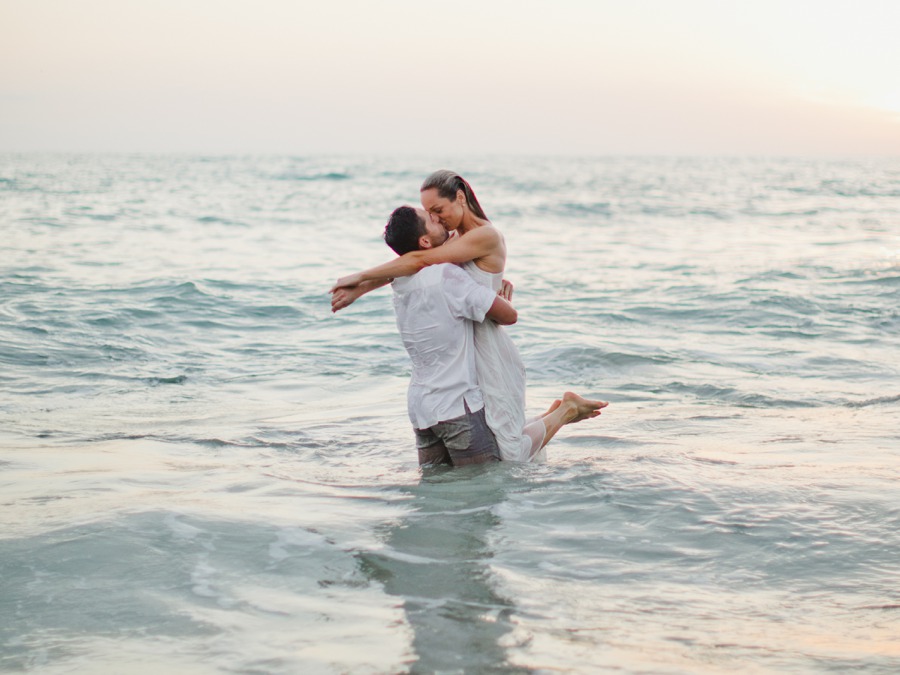 A man stands in the ocean holding a smiling woman in a white dress, both partly wet, with calm waves and a pale sky in the background—a beautiful moment captured during their Calafia Beach engagement photos.