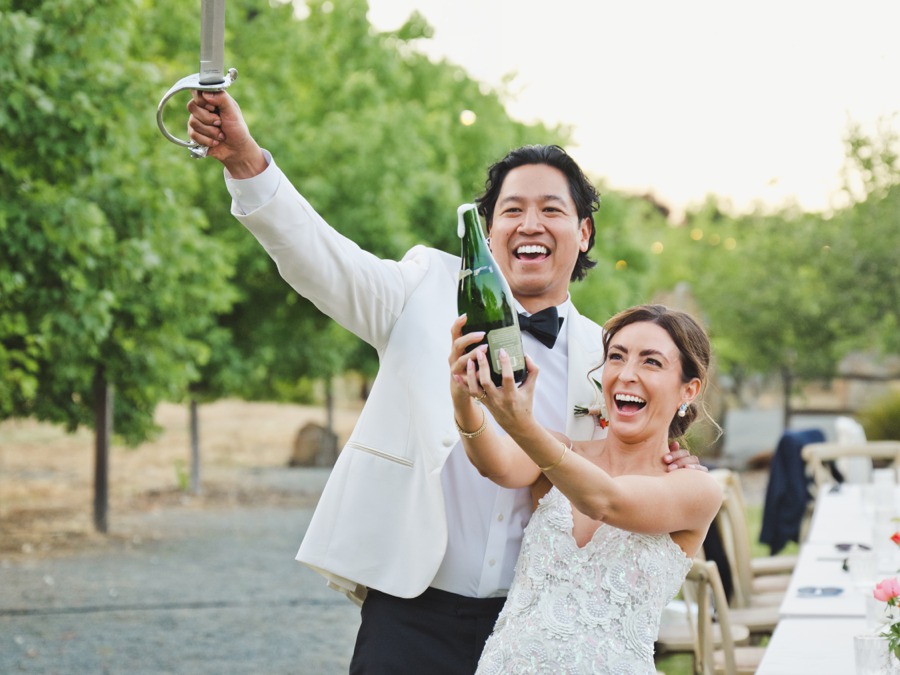 A couple in formal wedding attire smiles outdoors as the groom raises a sword and the bride holds a champagne bottle.
