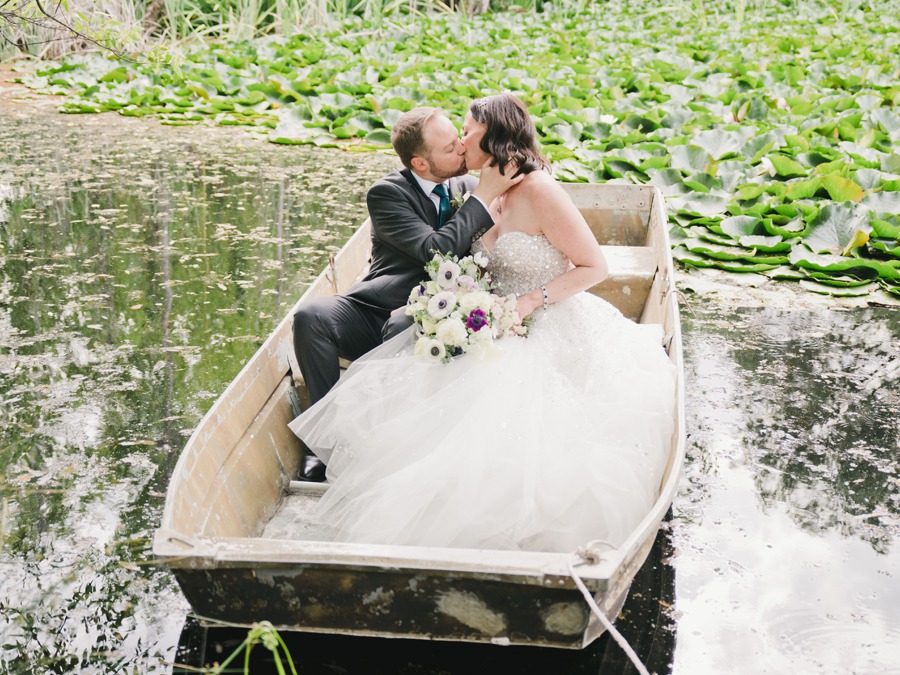 A bride and groom sit in a small boat on a pond surrounded by lily pads, sharing a kiss while the bride holds a bouquet.